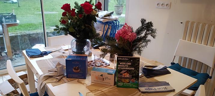 Sur la table de la cuisine, deux bouquets, l'un de roses rouges, l'autre d'une jacinthe rose. Ils ont tous les deux des petites branches de sapin.
Devant, une wonderbox, une boîte de Morphée (l'appareil pour méditer) et une autre boîte avec un petit appareil pour faire du yoga.