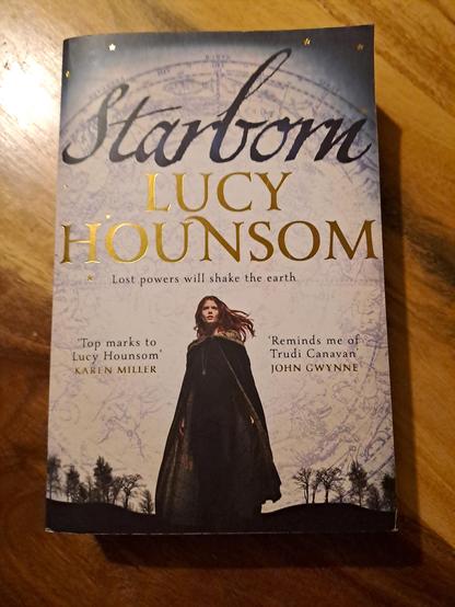The cover of the book.

A woman in a cloak stands looking forward, some trees in the distance behind her. A map of the zodiac is lightly drawn behind her.