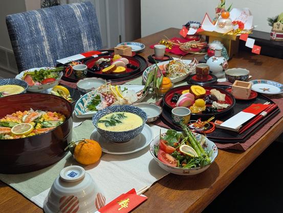 Japanese New Years dinner on a dining room table. The ones I can name are chirashi, veggie salad, chawan-mushi, sashimi with tsuma, sake (atsukan & reishu), two types of beans, wrapped kombu, and kamaboko. There are a few other foods that I'm not sure of the name of in English or Japanese. There are also Japanese New Years decorations at the top of the table.