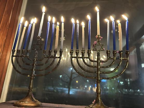 Two traditional brass chanukiyahs fully lit with a mixture of white and blue candles. The window is dark beyond.