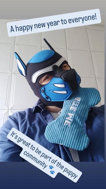 Pup Byte, ein Pup Player mit blau-weiß-schwarzem Hood, hat einen blauen Knochen aus Stoff mit der Aufschrift "Bite me" in der Schnauze. Darüber der Text: "A Happy new year everyone". Darunter steht: "it's great to be part of the puppy community ".