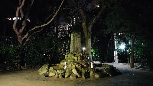 A park at nighttime. There is a clearing with a mound of earth which is surrounded by large stones. A tree is growing from the mound, and there is a stone marker of some kind with faint Japanese writing on it. There are more trees in the background, and the tops of distant buildings are just visible through them.