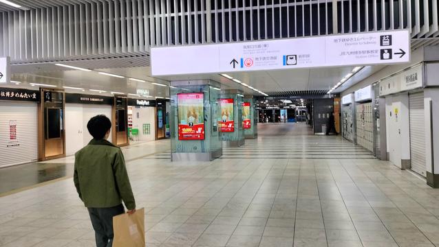 A Japanese train station at night. Ordinarily it is very crowded, but it is completely empty except for one man carrying a bag. The view is of a long corridor with a white tiled floor. Shops line both sides, all closed except for a convenience store. There are several illuminated advertising kiosks along the way. The man appears to be looking at a sign in Japanese and English that indicates where to go to find the elevator, the station office, etc. In the distance are doors open to the outside.