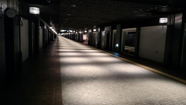 An underground shopping mall in Japan. It is a long corridor receding into the distance. It is completely dark except for some dim lighting along the corridor. Closed shops line either side of the corridor. The floor is made of flat, smooth, grey cobblestones.