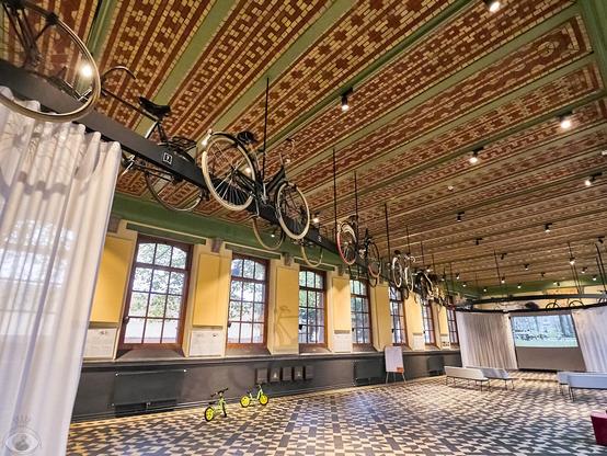 Old renovated building with brown/green mosaic tiled ceiling. Hanging from it, several (like a dozen) vintage bicycles. On the black and white mosaic tiled ground there is a couple child bicycles and at the end there is a screen playing a video about bicycles.
