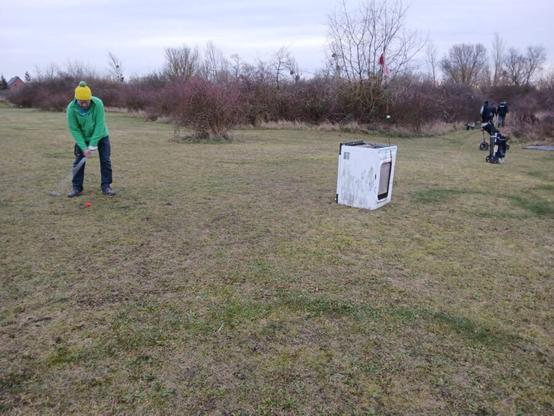 ein Crossgolf Spieler versucht aus einer sehr schrägen Position heraus in eine Frontlader Waschmaschine einzulochen