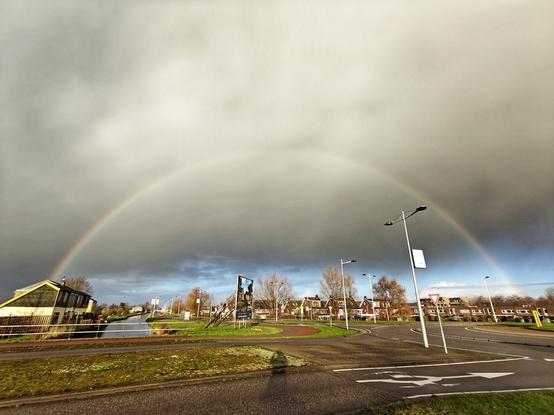Een regenboog met een weg op de voorgrond. Op de achtergrond, achter de wolken is blauwe lucht te zien.