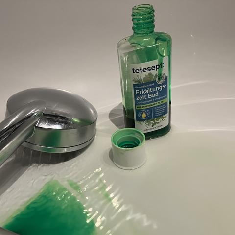 Inside of a bathtub, the shower hose the bottle and cap of “Tetesept Erkältungsbad” standing of a puddle of green gel (which is the content of the bottle, a mixture of essential/ethereal/volatile oils meant to loosen respiratory congestion when added to bathtub water).