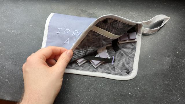 A hand lifts a corner of the case, revealing a grey-ish pattern inside fabric and a few keys of a small keyboard also held in white and lavender.