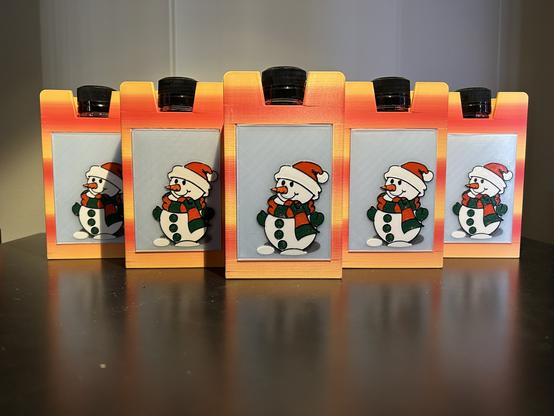 Five 3D printed boxes are lined up.  They have soft horizontal yellow and orange stripes.  Each has the cap of a flask protruding out of a notch in the top, and each has a six color printed picture of a snowman wearing a scarf and a Santa cap.