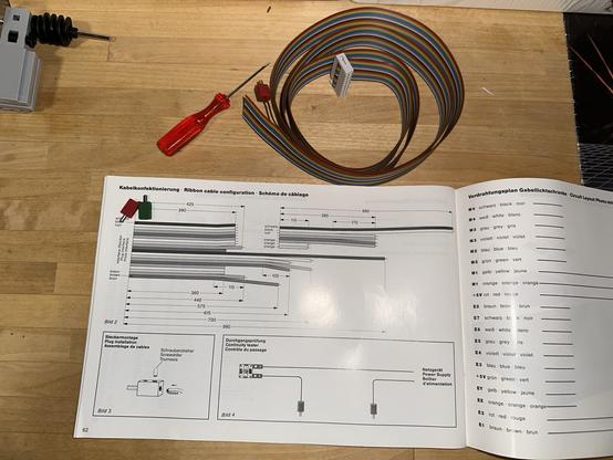 A ribbon cable with 20 colored wires, rolled up. A red screwdriver. Some small Fischertechnik plugs in red and green. A manual page showing the "ribbon cable configuration" with the precise lengths the wires must be trimmed to.