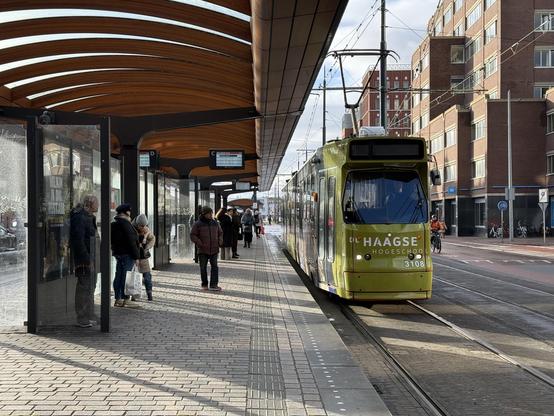 Photo of a green tram on the right and a platform with roof on the left. The roof is wood and wood colored. The tram is wrapped in green livery with an advertisement for “De Haagse Hogeschool”.