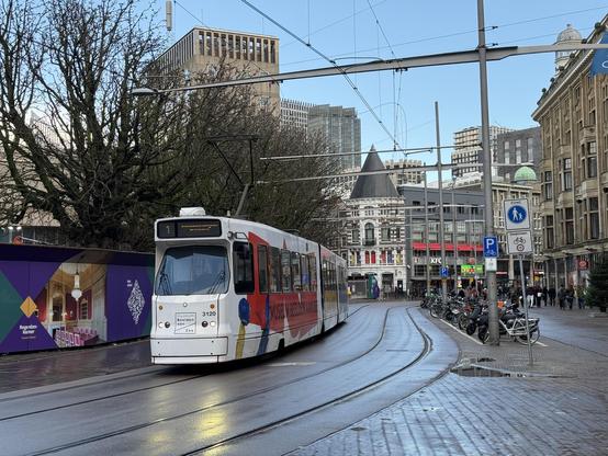 Photo of a white tram on the left. The tram track is in the middle and pedestrians on the right. On the right also some buildings.