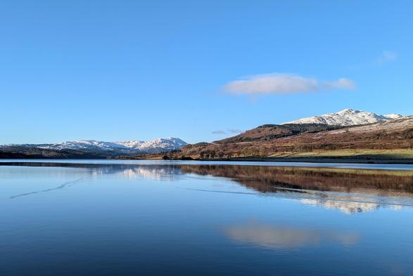 A view on a cold, sunny, clear day across a loch towards some snow-topped hills. There is a lower hill in front without snow, and two behind which are white with snow. The water of the loch reflects the hills, with a few ripples.