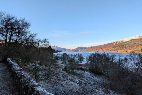 A view on a cold, sunny, clear day across a loch towards some snow-topped hills. In the foreground is a low stone wall, with a thick layer of frost on top. Below is a frosty field that goes down to the loch; and then across the water are the hills.