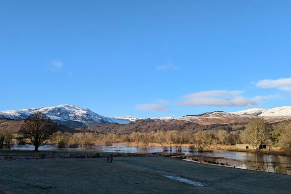 A view on a cold, sunny, clear day across a field, over a river in spate and across towards some snow-topped hills. There are a couple of people walking in the field.