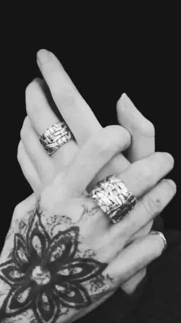 Bkack and white photo of two holding hands wearing woven silver rings on a black background