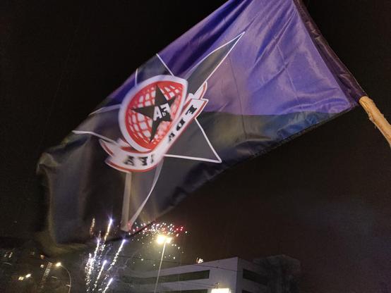 Photo of a waving purple-black (anarcha-feminist) flag on a wooden stick. The flag has a star and the logo of FDA-IFA in the middle (letters mirrored). In the background: black sky and exploding fireworks.