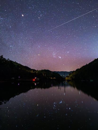 Eine helle Sternschnuppe zieht ihre Bahn über den Winterhimmel mit dem Sternbild Orion, das sich in einem See spiegelt