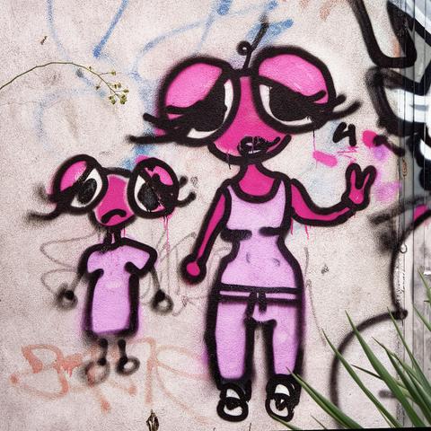 Graffiti mit zwei in rosa gehaltenen Figuren. Übergroße Augen. Etwas mäuseartig. Die kleinere Figur rechts, vielleicht ein Kind, schaut traurig mit hängenden Armen. Die größere Figur eher optimistisch mit zum V gespreizten Fingern der linken Hand.