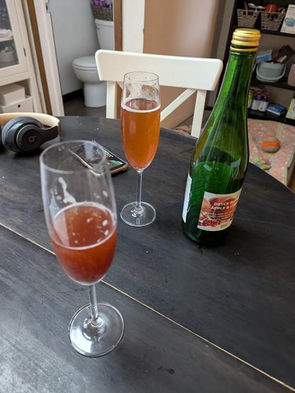 Picture of two champagne flutes with orange-red beverage in them, next to Ikea lingonberry soda bottle on a black table.