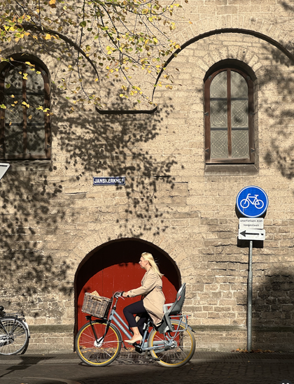 Person on a bike with a front basket and rear child seat. They are biking past a red door that is inset into an old stone building. A blue and white bike icon sign is to their right. The photo was taken in Utrecht, NL in 2024.