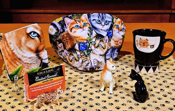 A cat-themed tea tableau with pawprint and catprint fabrics, handpainted mug, and antique porcelain figurines of cats. There is also a figurine of a Bengal cat, one of the noisiest of all felines. Appropriately enough, the tea is Constant Comment black tea from Bigelow.