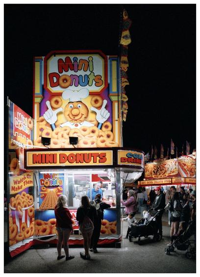 The image depicts a lively outdoor fair or carnival scene at night. Prominently featured is a colorful food stall selling mini donuts. The stall is vibrant with cartoonish designs, including a smiling character made out of a donut, wearing a chef’s hat, on the signage. Below the character, words that read “Mini Donuts” are prominently displayed. The stall offers a welcoming and cheerful atmosphere with lights and displays advertising mini donuts, as well as other items like "Cold Drinks." Several people, including adults and children, are either engaging with the vendor, ordering food, or are nearby observing and walking. The scene captures a typical festive and casual environment typical of a fair, emphasizing community enjoyment and casual dining.