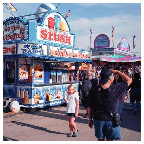 This image captures a lively scene at a fair or outdoor festival. It features a colorful food stand advertising "Fruit Flavored Slush," "Ice Cream Cones," "Sundaes," and "Ice Cold Water." The stand is decorated in vibrant blues and yellows, and it's bustling with activity around it.

There are several people in the image, including a young girl standing in the foreground, almost center frame, who appears to be eating or holding something close to her face. To her left, an adult male is seen from behind, wearing a black T-shirt and a camera around his neck, and he's shading his eyes with his hand.   The triangular space created by his arm and head is occupied by the face of a woman walking towards them.

The overall atmosphere suggested by the image is vibrant and festive, typical of a fair with food stands and possibly rides, as hinted by more structures in the background that resemble part of an amusement ride setup. The time of day seems to be during daylight hours, under clear skies, contributing to the bright and cheerful setting.