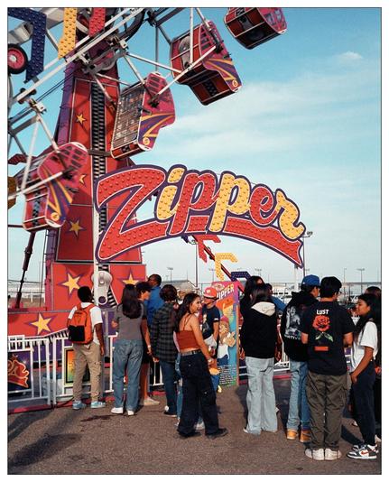 This image shows a scene at a fair or carnival featuring the "Zipper" ride, which is a popular amusement ride known for its rotating cages and spinning motion. There are several people in line waiting for their turn to ride. The atmosphere appears lively and recreational, with the large, colorful sign of the ride in the forefront adding to the festive feel. The sky is clear indicating it's likely a pleasant day for outdoor activities.