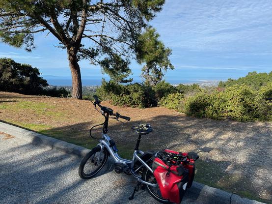 Mid-afternoon sunshine above the blue waters of the Monterey Bay. A Tern NBD bicycle with red panniers is parked along the edge of the road.