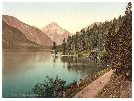 The image depicts a serene landscape featuring a tranquil lake with clear blue waters reflecting the surrounding greenery and mountains. The foreground shows a winding dirt path that leads towards the water's edge, bordered by dense trees on one side, suggesting it is likely late spring or summer when vegetation is lush. In the background, towering mountain peaks rise majestically against a soft-hued sky transitioning from pale blue to gentle shades of orange and pink at the horizon line. The overall atmosphere conveyed by this image evokes feelings of peacefulness and natural beauty.

Additional information about this particular photograph indicates that it was taken in Plansee, Tyrol region within Austro-Hungary during roughly 1890-1900s period. It is part of a collection known as "Views of the Austro-Hungarian Empire," which suggests its historical significance and serves to provide context for viewers interested in exploring this specific time frame and geographical location through photography.

The photograph's vintage appearance, with noticeable coloration variations across different elements like water, trees, sky, etc., is indicative of early photographic processes. These inconsistencies might be due to the aging of prints or intentional artistic choices made during its production era. The image has been identified by a reference number (17150) and associated text that implies it may have historical importance f [...]