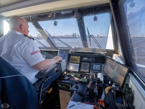 Wide angle shot from the right side. The bridge of the waterbus is quite small with only a seat for the captain and barely any room to stand for a second person. Several screens with all kinds of data (a digital compass, meters displaying the motors, etc.) In front on a board next to the captain a pair of binoculars, cellphone, sun glasses, etc and also a banana. The harbour of Antwerp can be seen from the window.