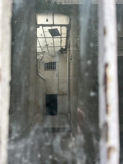 Looking through a dusty windowpane framed slightly off centre by metal bars painted white into an abandoned neoclassical house. The narrow courtyard would have once been open to the sky but has been roofed over in framed panes of glass in two different sizes, larger to front and not quite aligned. There’s a barred window high up in the back wall, half draped in white cloth and a dark doorway underneath. Everything is white or shadow, though this is not a black-and-white photograph. A larger doorway to the right, two levels in the tiled courtyard. There’s image is barely overlaid with the reflection of the apartment building across the street.
