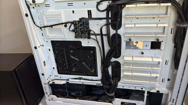 Same state of the workstation, other side (with panel removed). A central small black PCB is visible, with straight, black, folded, wires, zip-tied into rigid lines. There are black grommets that the black wires pass through in the otherwise white case, which further mask their existence. I hate seeing wires. The PCB is the independent fan controller of the case, which is a "be quiet! 802". A column of white bays rises up on the right side, while underneath, the power supply of the system, also black, with black, webbing-wrapped wires, is shrouded in darkness.
