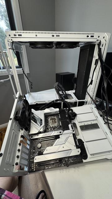 The L-shaped metal bracket which holds the motherboard, rear sockets, and the rear case fan, now rests in my hand, still connected by a few power cables at the base. This truly is like surgery. The near-bare skeleton frame of the computer resembles bones. Two fans are still in place at the underside of the top. They are also black, as is the 420-size radiator, which takes up the entire front wall of the tower (fans are on the other side of the radiator). The room is grey with white trims and it is comfortably bright daylight.