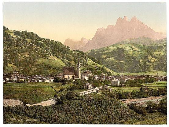 A picturesque vintage photograph showcasing a quaint village nestled amidst rolling hills and lush greenery. The backdrop features towering, jagged mountains under a soft, pastel-hued sky, suggesting either dawn or dusk lighting conditions. Notable architectural elements include a church with a prominent steeple, surrounded by traditional buildings that create a harmonious blend of human habitation within the natural landscape. This idyllic scene captures an era where nature and rural life coexisted in peaceful existence during Austro-Hungarian Empire's period between approximately 1890 to circa 1900.