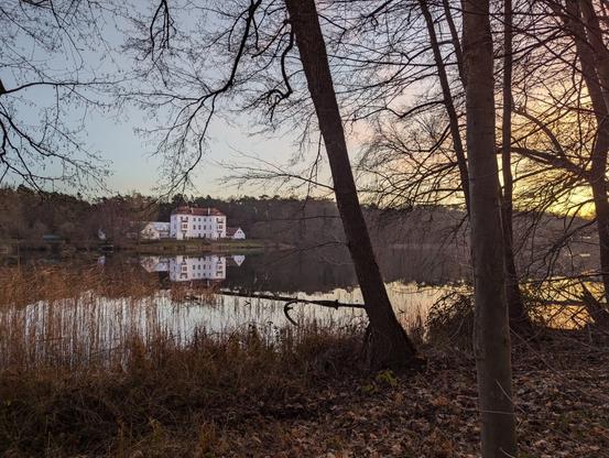 Across the lake, a small castle is visible under pastel colours of the winter afternoon sky. The sky also gives colour to the water, all trees are leafless.