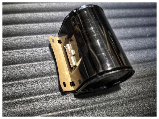 A Rapid film canister on a grey background. Sticking out of the cassette is the end of a roll of film.
