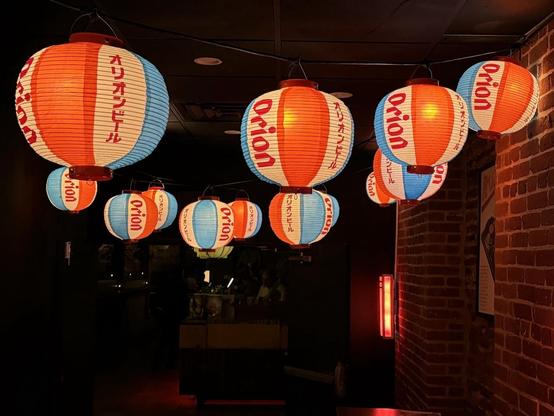 Many round paper lanterns with blue, white and orange-red stripes.  Brick wall on right.  Dark bar beyond.