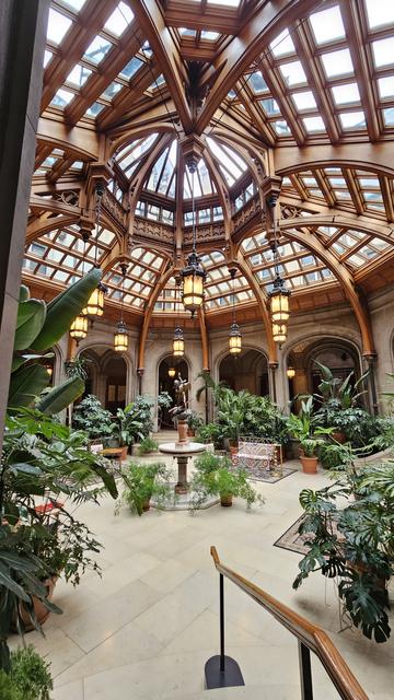 A large room of chateauesque architecture, with meter-long off-white tile floor and decorated with many green potted plants, some large and tropical. The concave top is covered with glass panes between thick light brown wood accents and supports. A center section extends even higher with long vertical glass panes. This section has ornate carved wooden decorations and details.

There is a round table in the center with a tall vase, and a small table and chair visible and rugs amongst the greenery along the sides. Eight large metal lanterns with yellow panes hang from the ceiling in a circular pattern, reflecting the shape of the room. The back wall is visible and is made up of stone arches that allow access to a hallway.