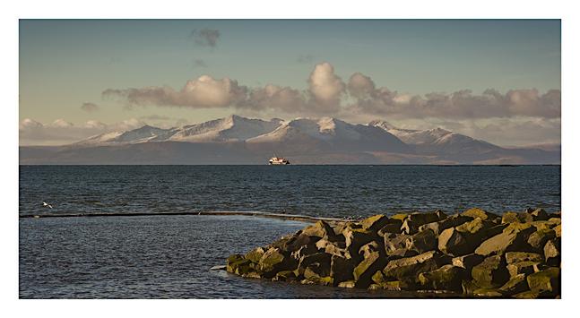 There are some rocks in the sea in the foreground. In the distance (very small) is the CalMac ferry heading for Brodick. The hills of Arran dominate the background and they have a light covering of snow.