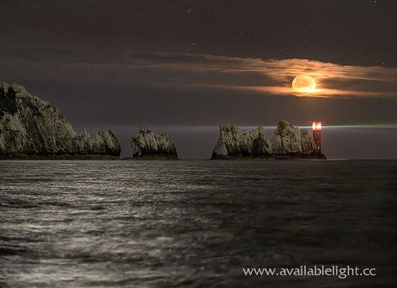 A bright moon, in a very dark sky over a lighthouse standing at the end of some tall chalk stacks. An inky sea is reflecting light in the sky. A beam of straight thin light is being transmitted across the picture from the lighthouse.