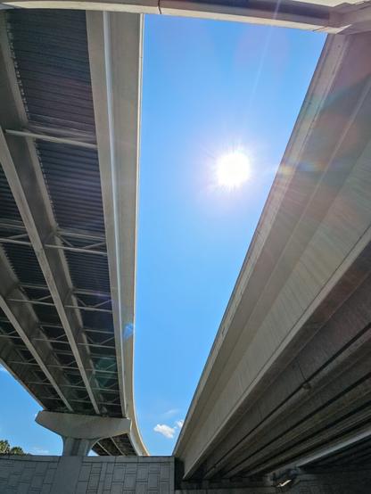 Mid day sun shines bright through an opening up above where different lanes of a bridge overpass curve around in different directions.