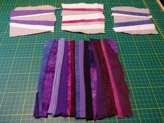 Four pieces of fabric made from strips of other pieces of fabric sewed together. The three at the top are made from solid shades of plain cotton: grey, white, and a colour (purple, pink, and blue respectively). Some of the strips in these are almost triangular, creating a rather jaunty effect overall. The piece at the bottom is larger, and the strips are generally more even in width. These are mainly in shades of purple, with some blue, black, and dark pink. The fabrics are more varied: cotton, linen, double gauze, shiny polyester, and some are patterned and some plain.