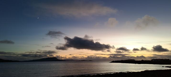 Looking southwest from Seil across Easdale Bay, there is a bright afterglow beyond some incoming clouds. As the sky graduates to a dark evening blue, the moon, planets and stars become visible. To the left of the image, just above and right of the crescent moon, is Venus.