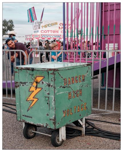 The image shows a scene from a fair or carnival atmosphere. In the foreground, there is a prominently featured weathered green cart with a "Danger High Voltage" warning sign painted on it along with a lightning bolt symbol. This cart appears to be connected to heavy cables, likely indicating it is part of an electrical setup for the fair.

In the background, multiple fair booths and signs can be seen, including those advertising "POPCORN", "COTTON CANDY", and "APP (APPLES)". There are several people visible in the scene, including a young child inside a fenced ride area looking playful or curious, and an adult man looking towards this child, likely implying a guardian watching over the child. The crowd and setting suggest a lively public event with typical fair food and attractions.