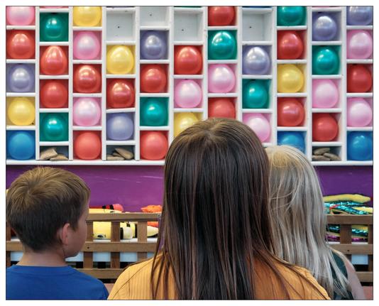 The image shows the back of three children looking at a wall-mounted shelving unit filled with colorful balloons arranged in neat rows within individual compartments. Each shelf has multiple square compartments, each containing a single balloon in various bright colors such as red, blue, yellow, pink, and green. Below the shelves, there appears to be a table with various prizes etc.  This is a dart and balloon game at the midway of a local carnival or fair.