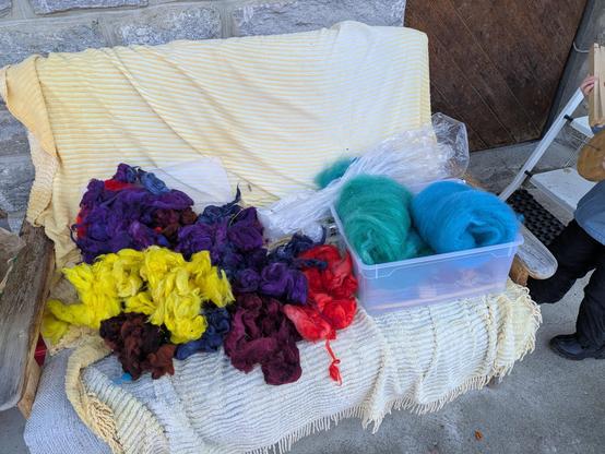 gefärbte wollflocken in verschiedenen Farben auf einem sofa. daneben zwei kardierte vliese in türkis und eisblau.