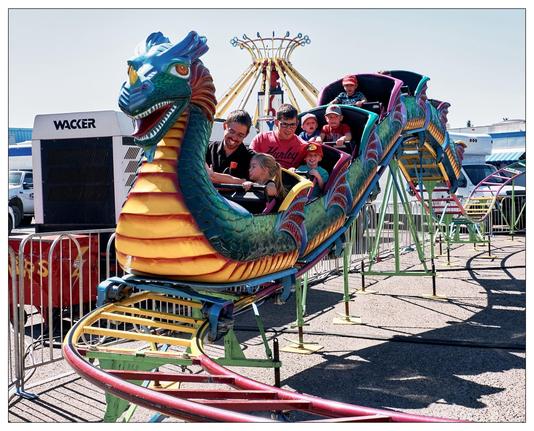 This image shows a group of people enjoying a ride on a dragon-themed roller coaster at a fair or amusement park. The front car of the roller coaster is designed to look like a vibrant, colorful dragon. The people on the ride are showing various expressions of excitement and joy. In the background, there are other amusement rides and equipment, which suggests this photo is taken in a lively outdoor setting, typical of a fair or carnival.